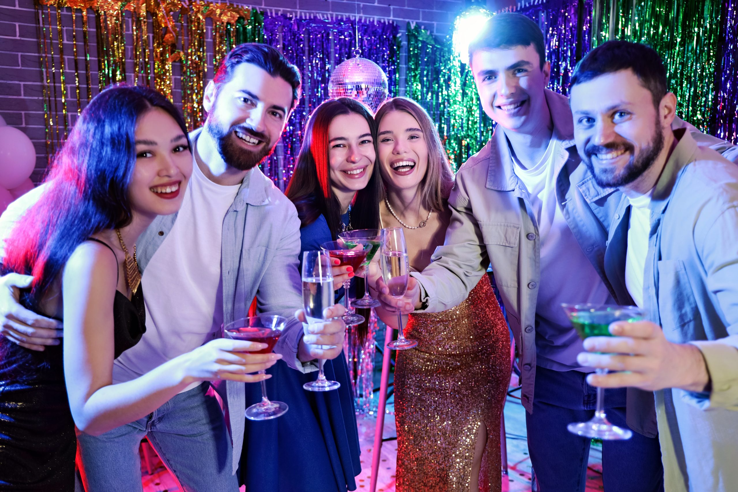 Six young adults dressed festively, smiling and holding drinks at a colourful party with tinsel, balloons, and a disco ball in the background—celebrating as if they’ve discovered all the exciting cruise lines and attractions awaiting them on their next adventure.