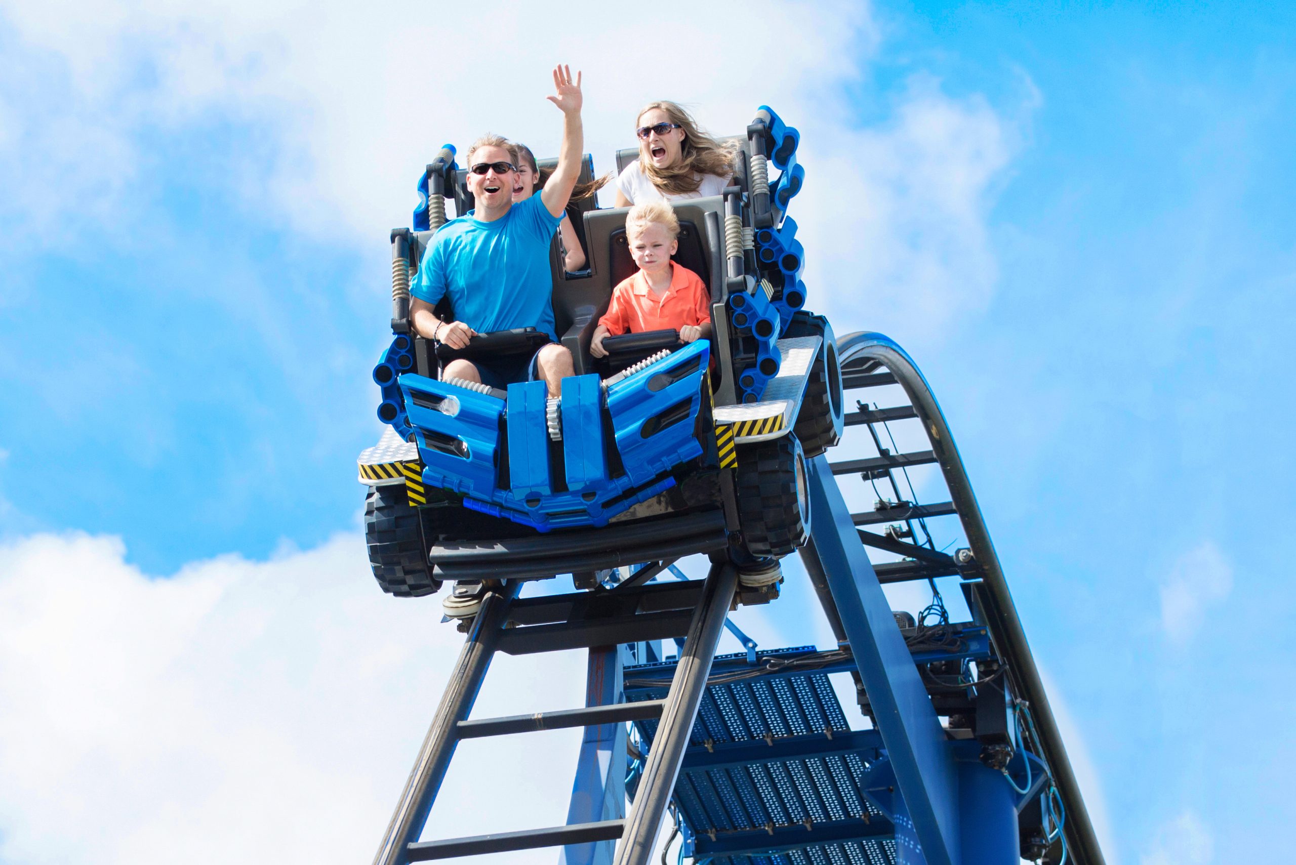 A family of three rides a blue rollercoaster, smiling and waving as they descend a steep drop against a bright blue sky with scattered clouds, showcasing the excitement found in top cruise lines and attractions.
