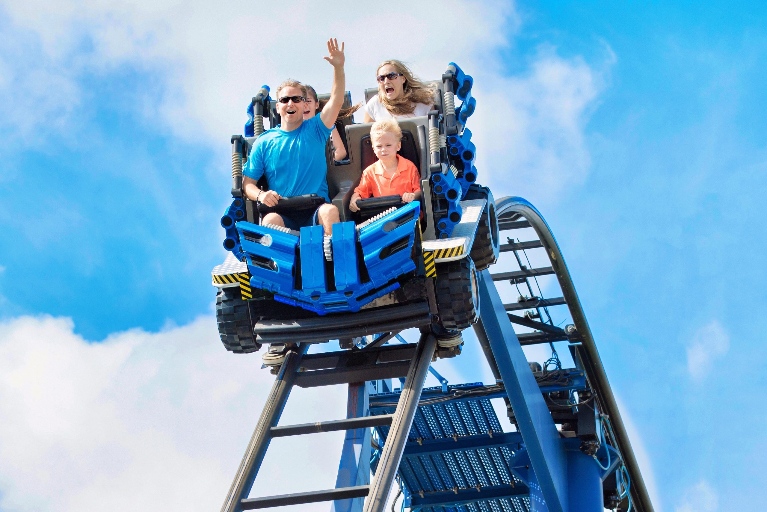 A family of three, two adults and a child, ride a blue rollercoaster car down a steep track—a thrilling attraction reminiscent of the excitement found on cruise liners—with bright blue sky and clouds in the background. The adults appear excited, while the child looks serious.