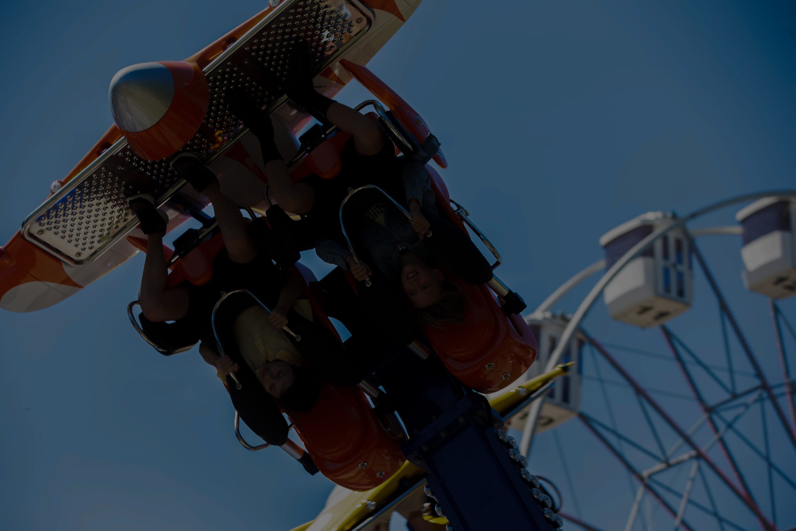 Two people ride an upside-down amusement park ride, smiling and holding onto safety bars, with a Ferris wheel and other attractions visible in the background against a clear blue sky—capturing the thrill often found on popular cruise liners.