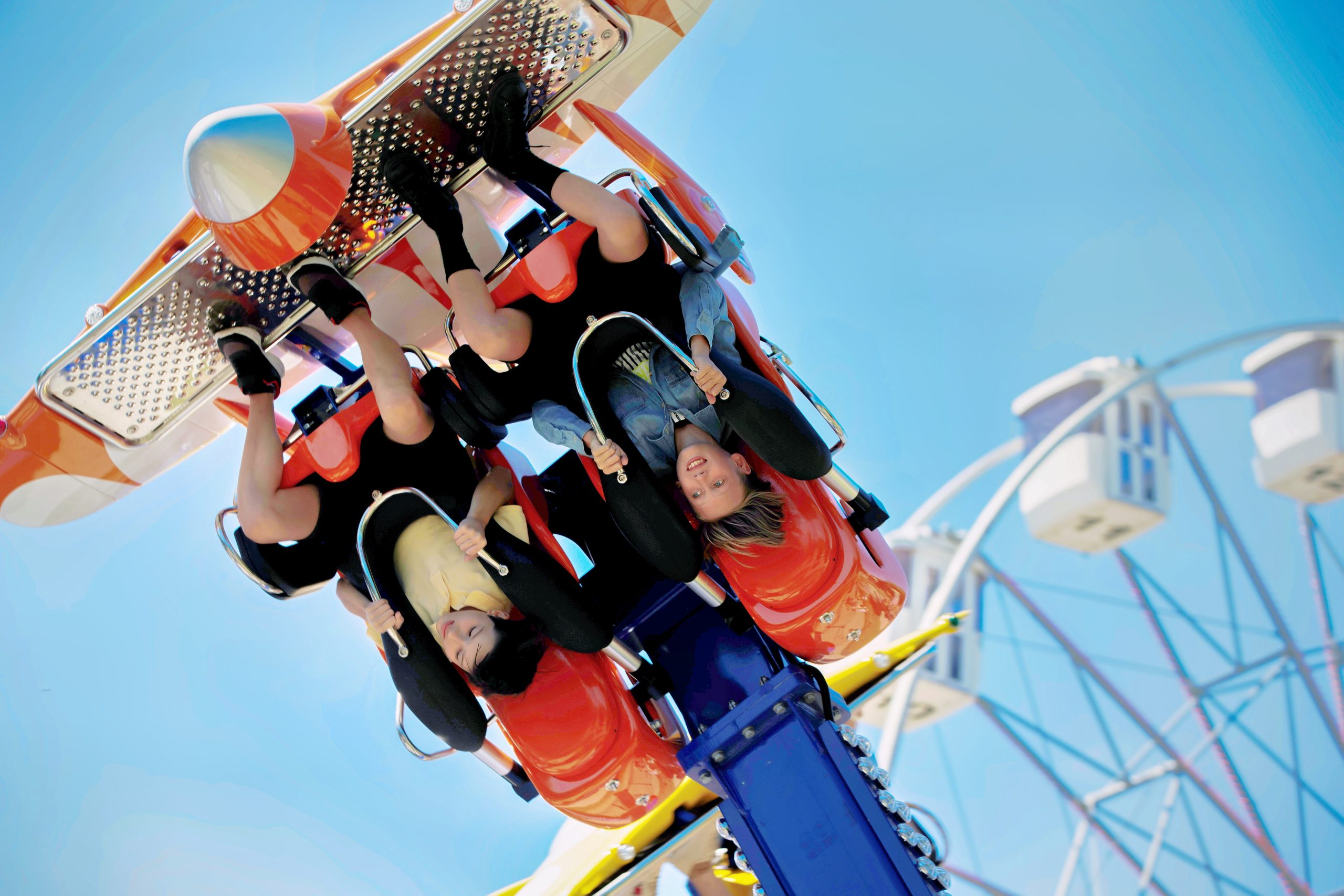 Two people are upside down on an amusement park ride, smiling and gripping safety bars, with a blue sky and a Ferris wheel blurred in the background—a thrilling moment that rivals the excitement found on popular cruise liners and attractions.