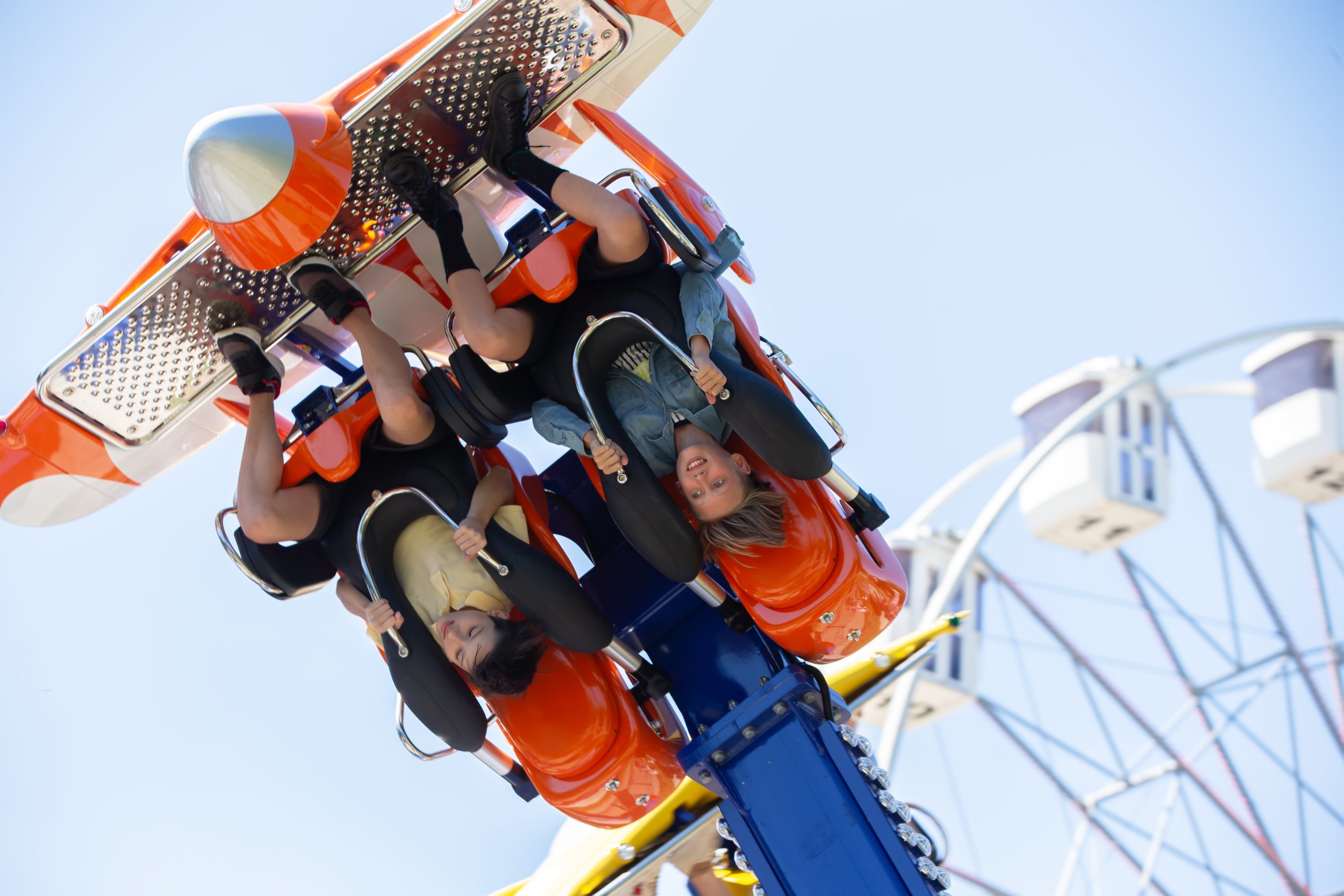 Two people are upside down on an orange amusement park ride, smiling and holding on to the restraints, with part of a Ferris wheel—one of the park’s many exhilarating attractions—visible in the blurred background against a blue sky, creating a scene reminiscent of the thrills found on major cruise liners and attractions around the world.