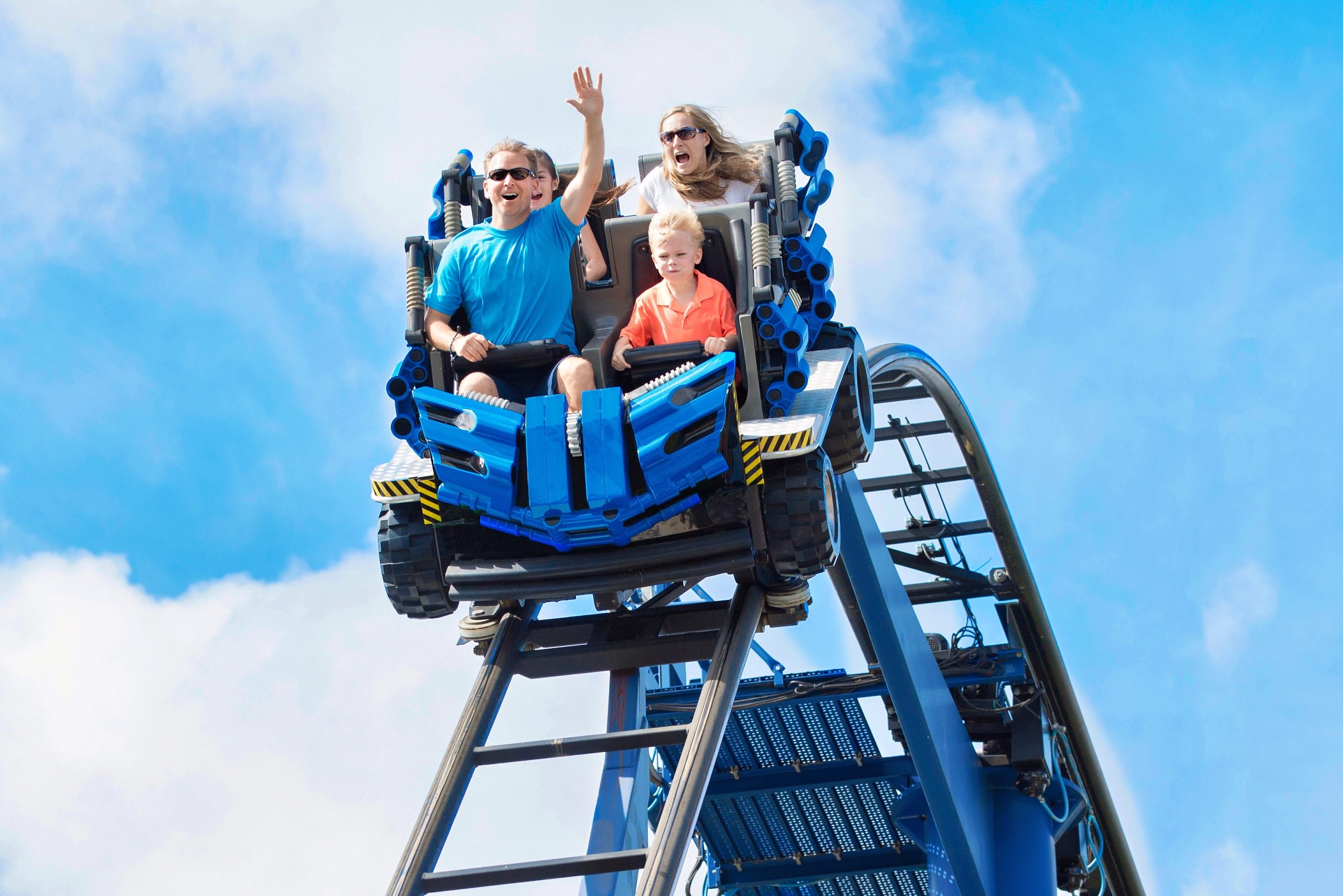 Three people, two adults and a child, ride a blue roller coaster—a thrilling highlight among various cruise lines and attractions. The adults look excited, with one raising a hand, whilst the child looks serious. The sky is bright and blue with some clouds in the background.