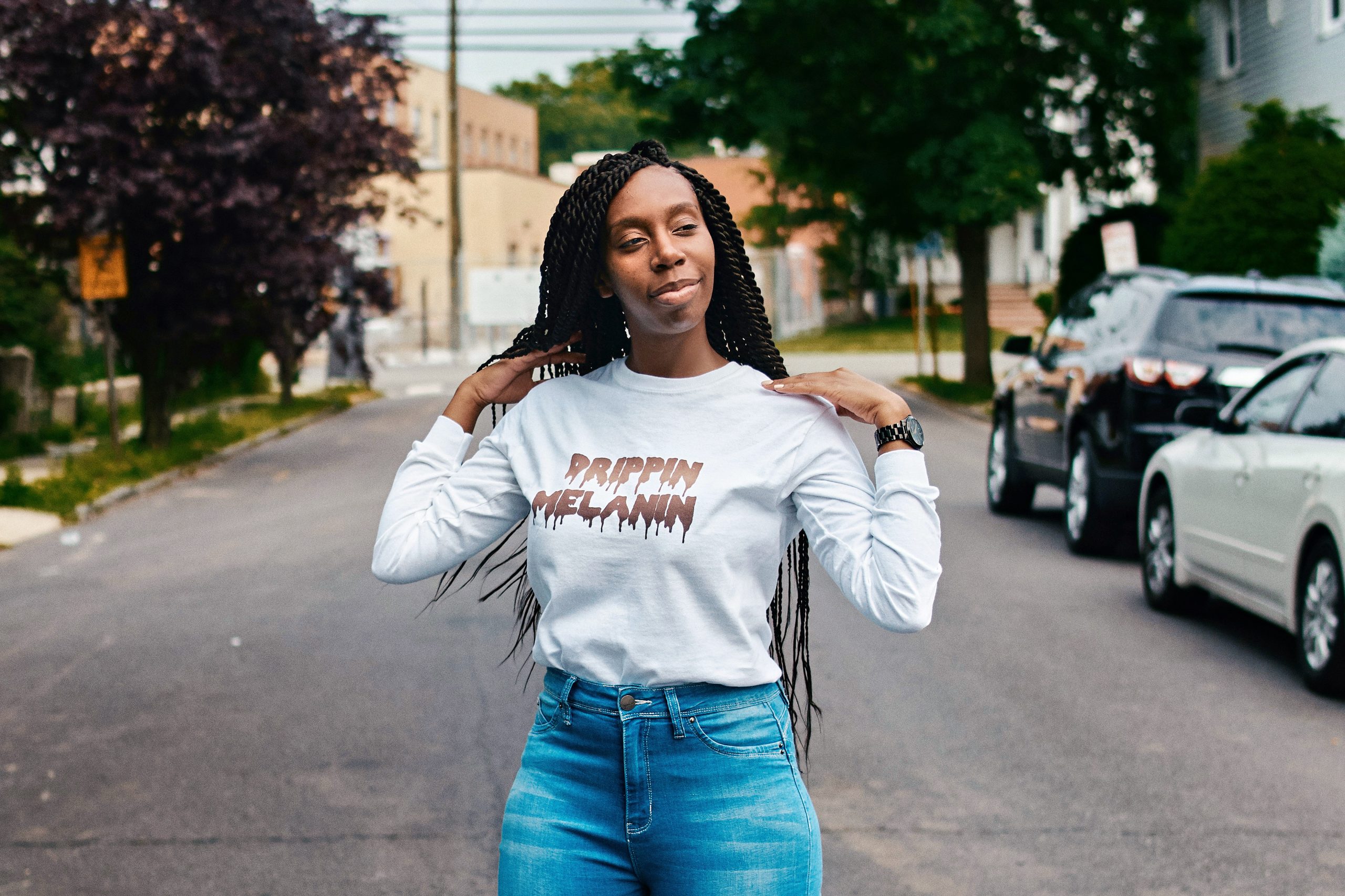 A woman with long plaits stands confidently in the middle of a street, as if she’s ready to take on any challenge—even in an API Playground—wearing a white long-sleeved shirt and blue jeans, with cars and trees forming the backdrop.