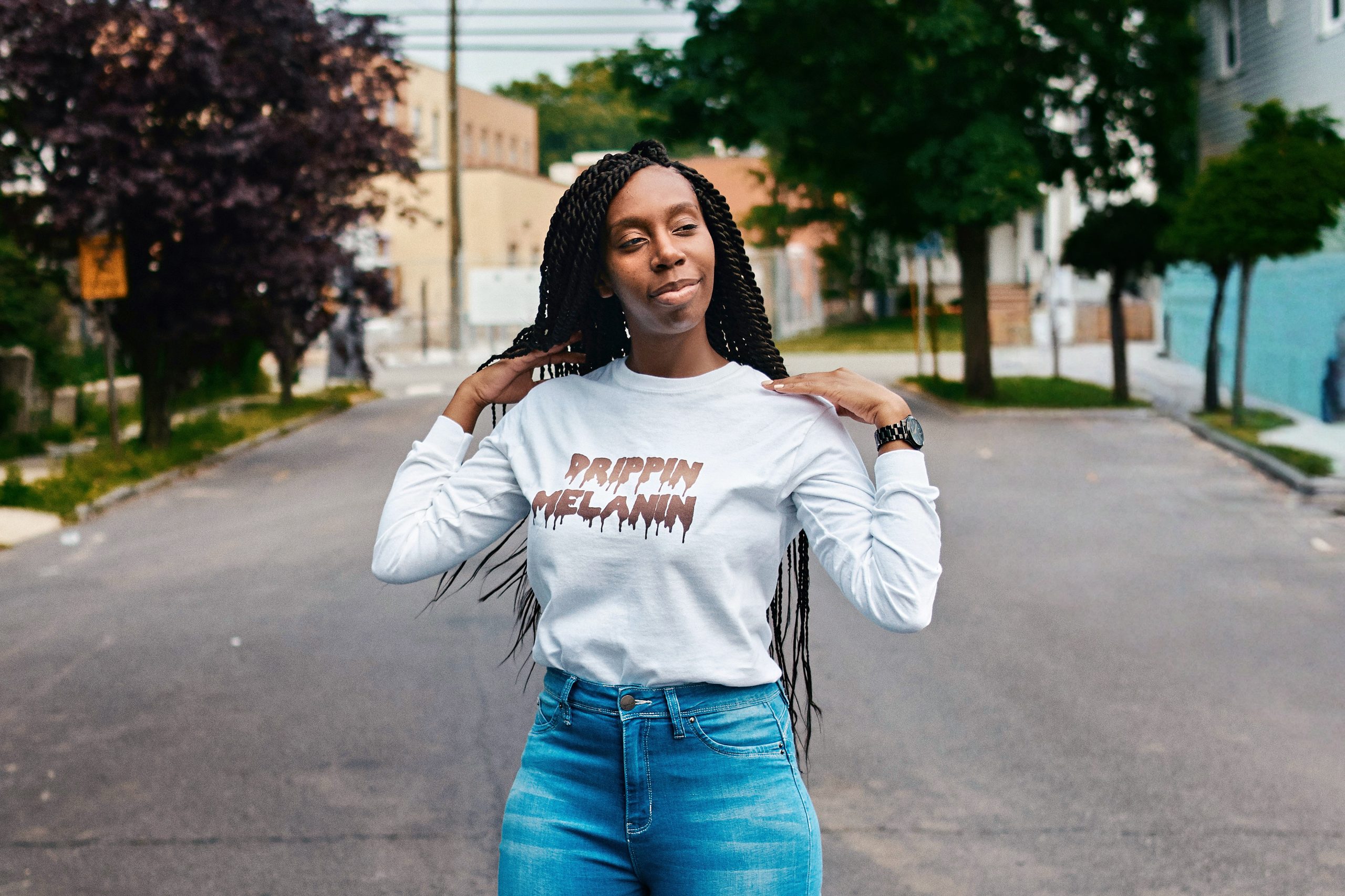 A woman with long plaits stands confidently in the middle of a quiet street, wearing a white long-sleeved shirt and blue jeans, as if she were contemplating her next big idea for the API Playground, with trees and buildings in the background.