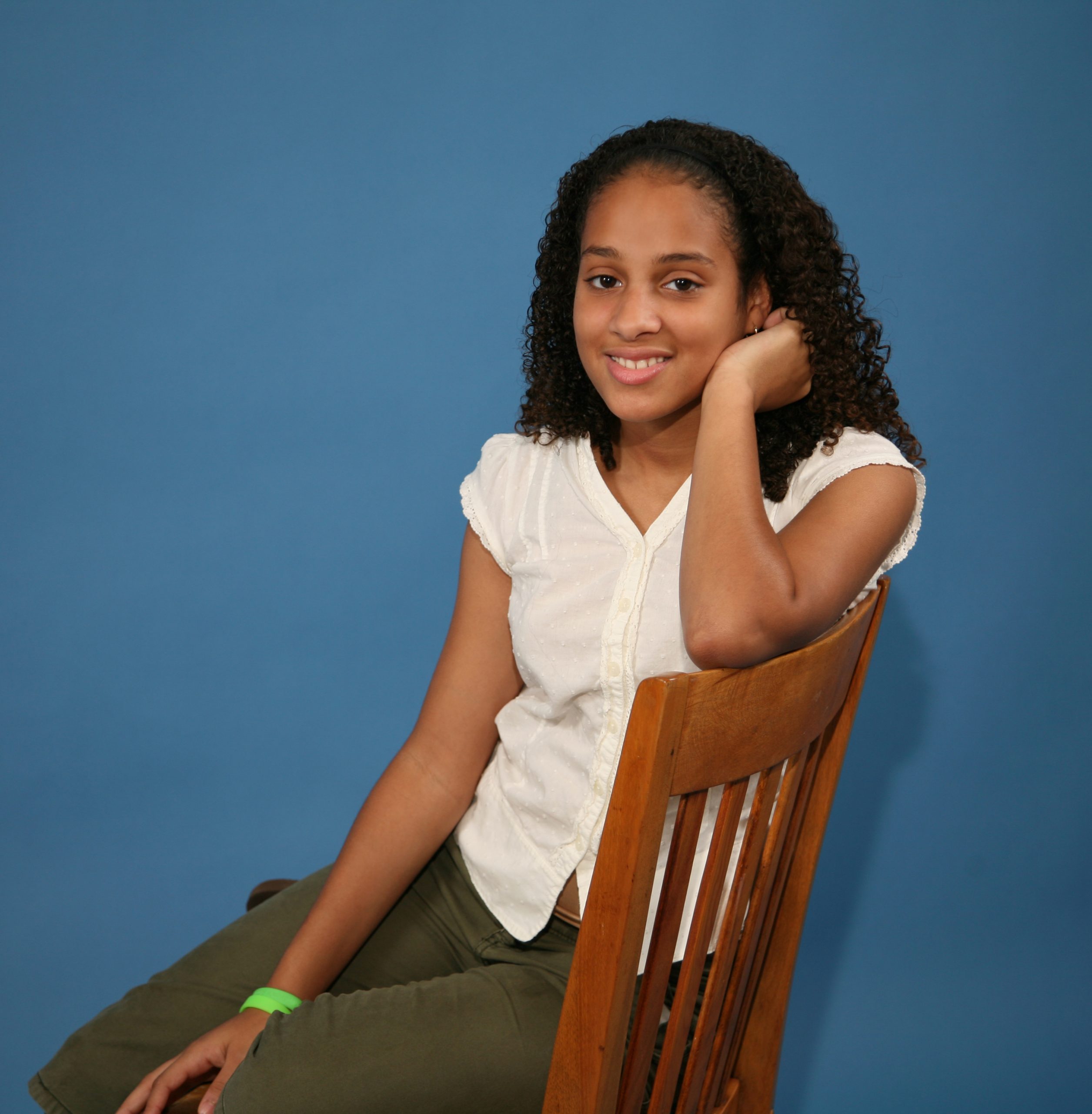 A young woman with curly hair, wearing a white blouse and green trousers, sits sideways on a wooden chair, smiling gently in front of a plain blue background, evoking the calm and approachable atmosphere of an API Playground.