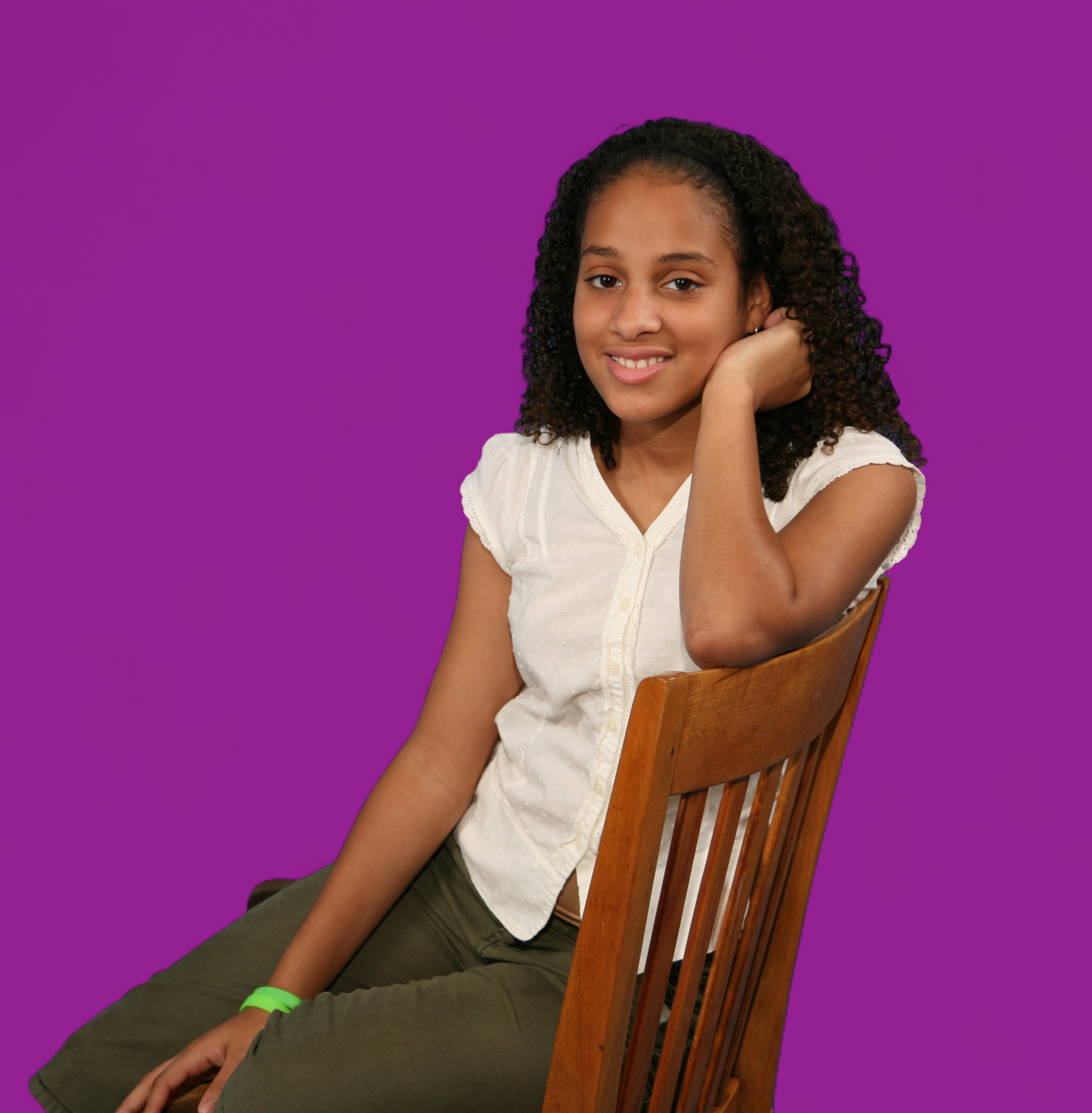 A young woman with curly hair sits sideways on a wooden chair, smiling and resting her head on her hand as if she’s just discovered something delightful in the API Playground, all set against a solid purple background.