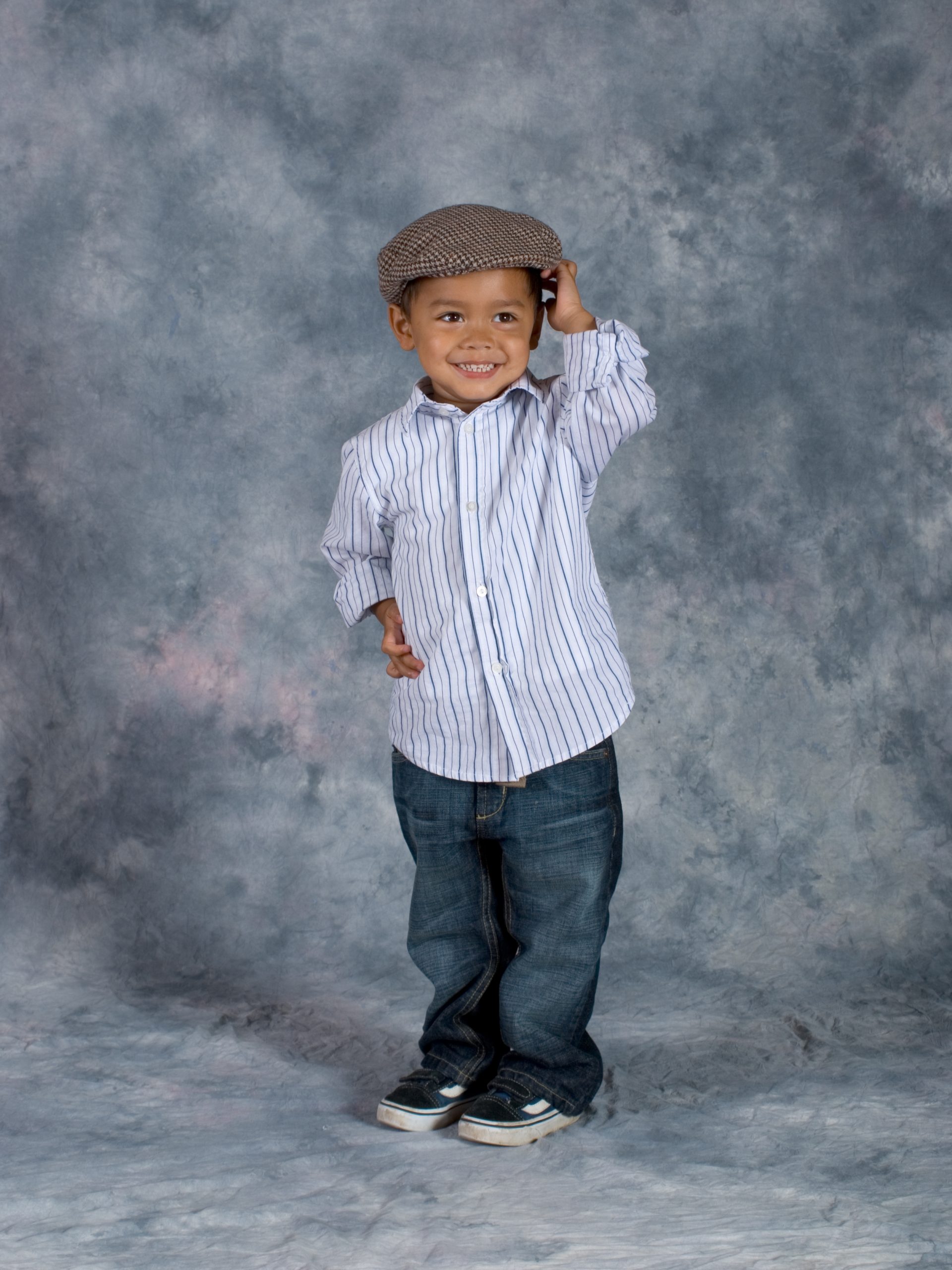Smiling young child poses in front of a grey marbled studio backdrop, wearing a striped button-up shirt, blue jeans, black shoes, and a brown flat cap, with one hand holding the hat—capturing a look as playful and creative as an afternoon spent exploring the API Playground.