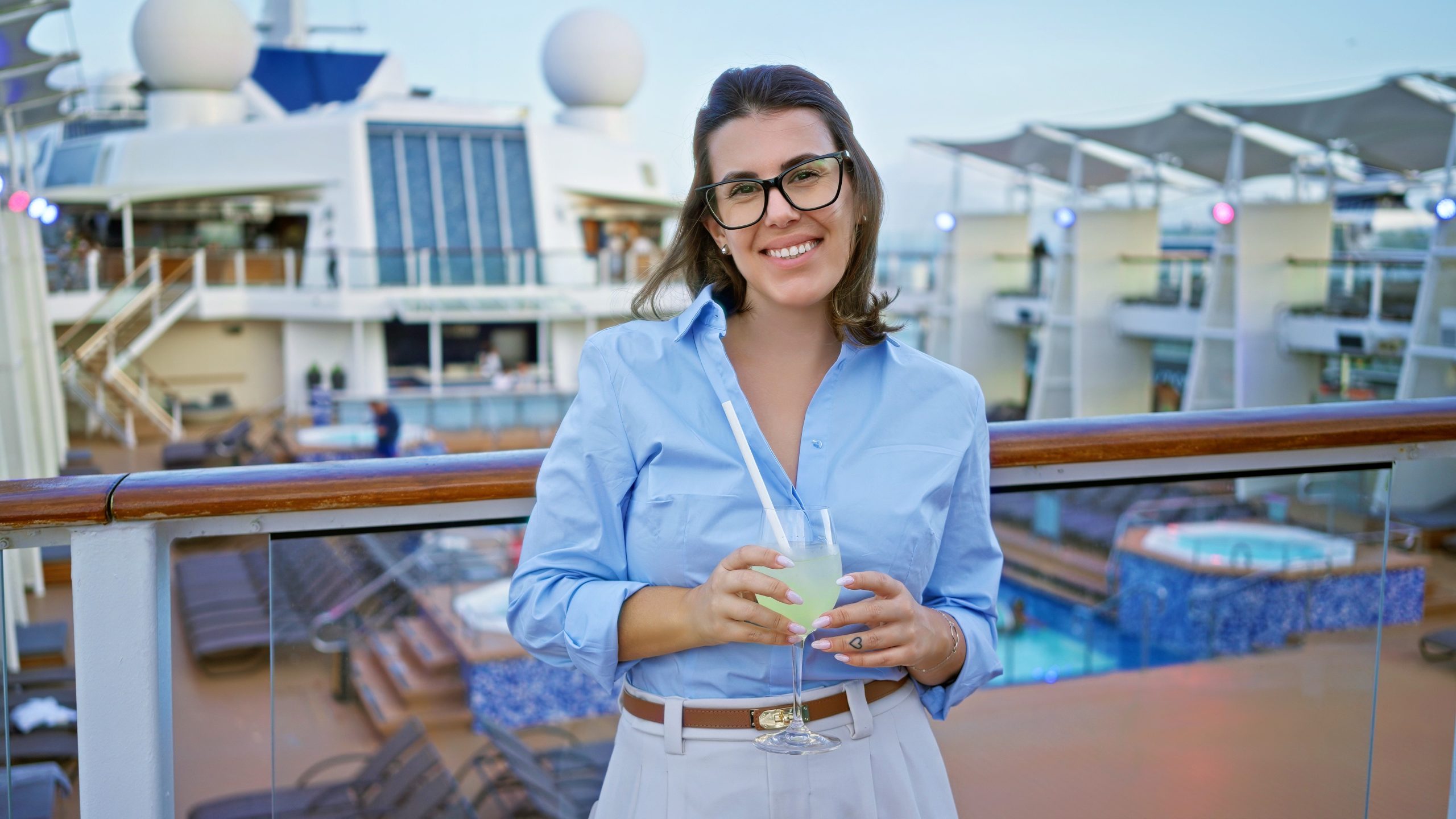 A woman in glasses smiles whilst holding a drink on a cruise ship deck, with pools and loungers visible in the background, enjoying the relaxing atmosphere that feels like an API Playground for leisure and adventure.