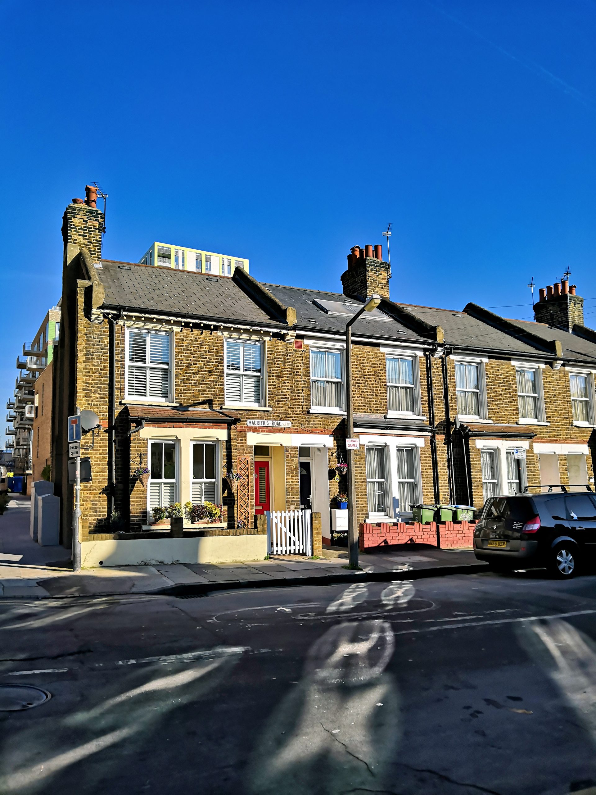 A row of brick terraced houses with white trim and chimneys stands under a clear blue sky, resembling a scene you might explore in an interactive API Playground. A black car is parked on the street in front, where shadows and reflections are visible on the road.
