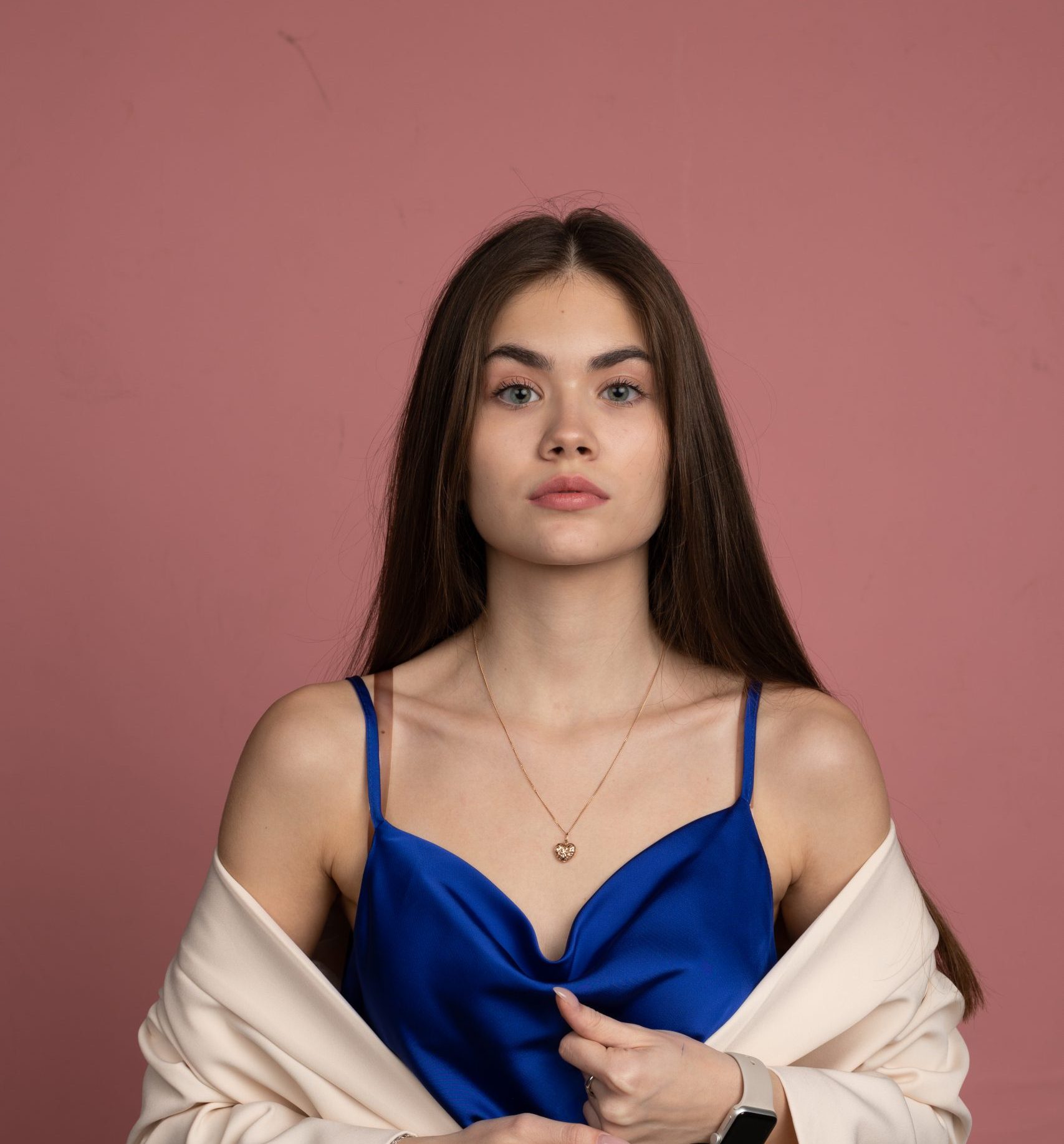 A woman with long brown hair wears a cream blazer over a blue satin top, holding the blazer’s lapels. She has a neutral expression and stands against a plain pink background. She wears a necklace and a smartwatch, blending modern style with contemporary tech—perfect for someone ready to explore the possibilities of an API Playground.
