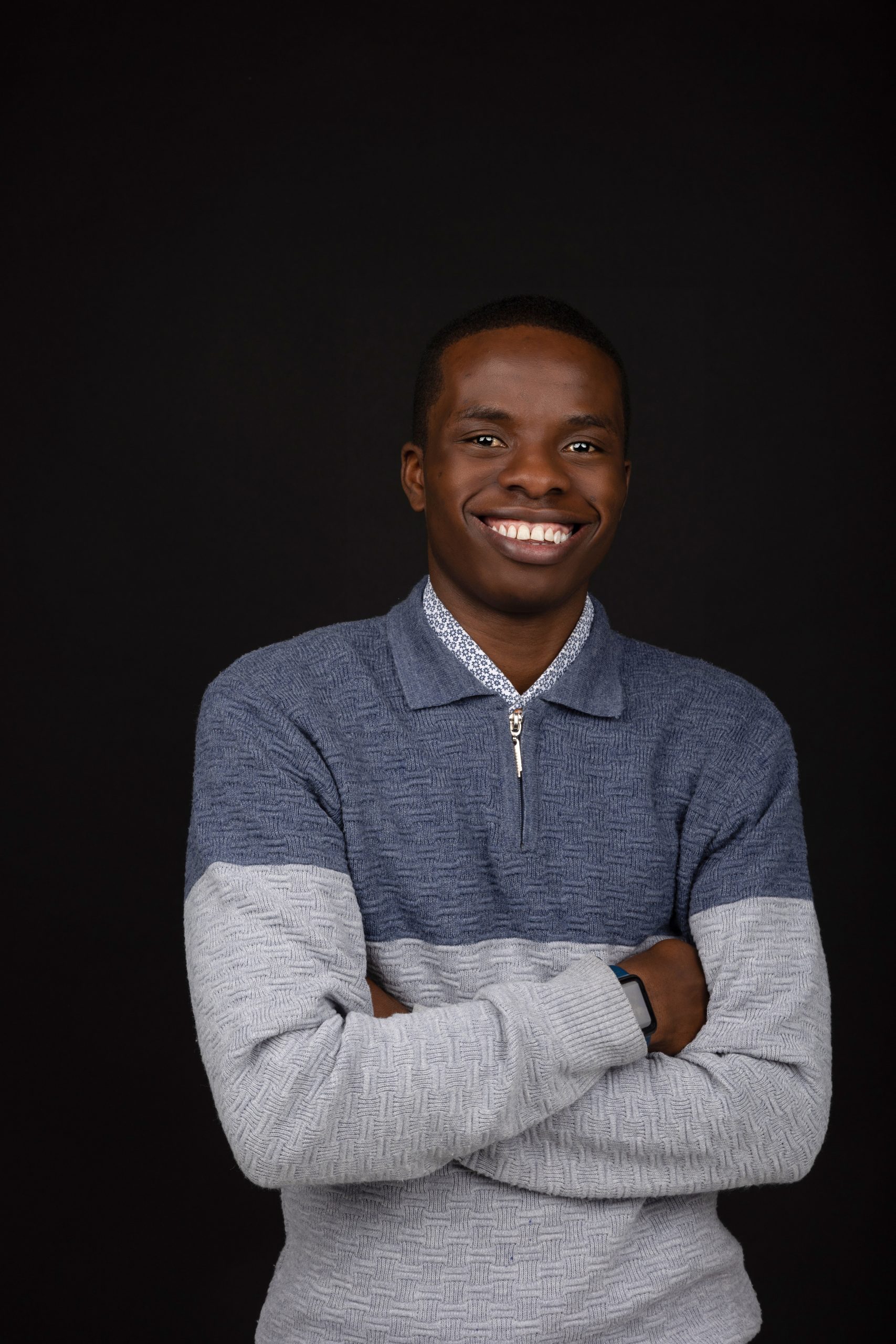 A person wearing a blue and grey jumper stands against a black background, smiling with arms folded—ready to showcase their confidence and creative spirit, just like someone stepping into a print lab for inspiration.