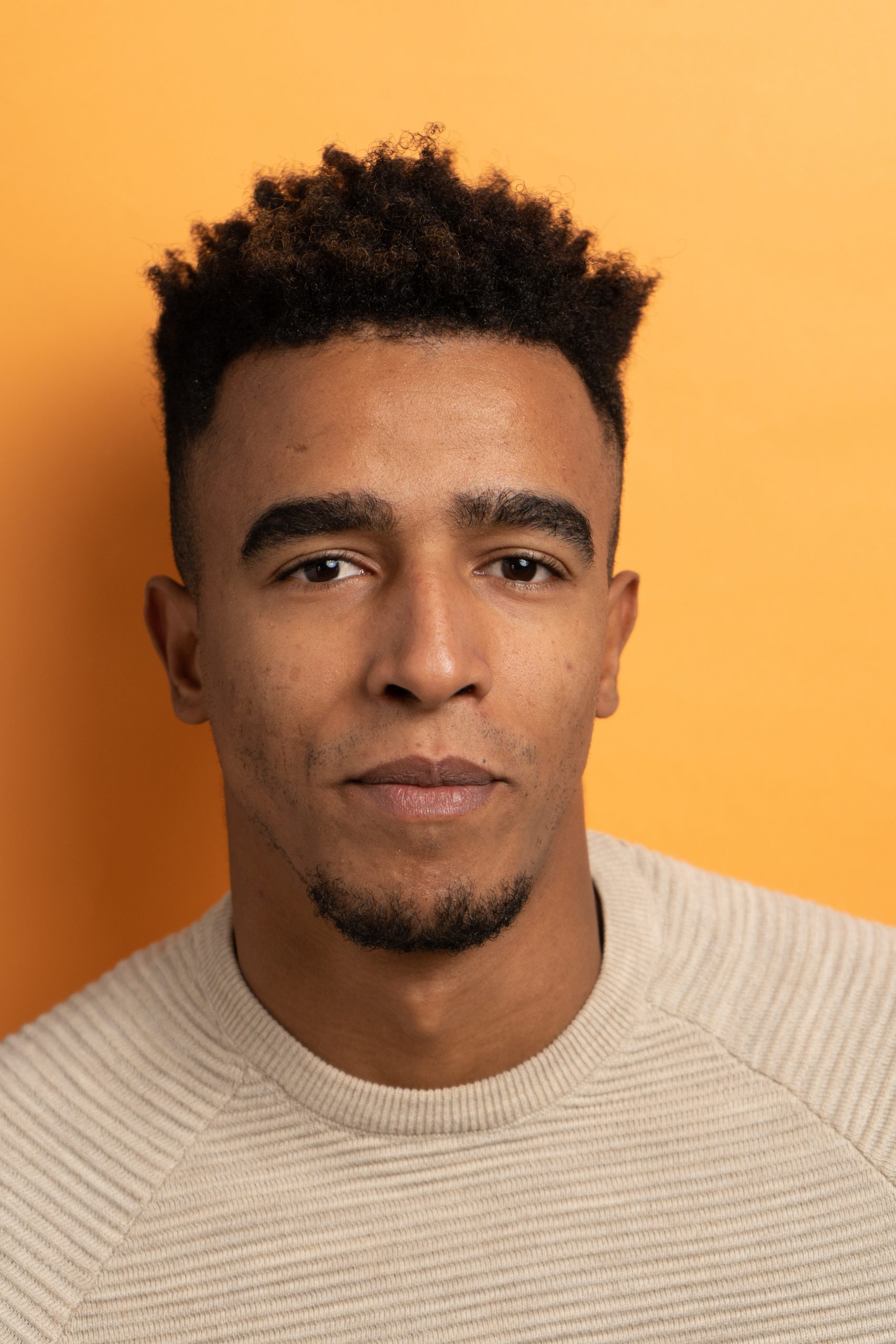 A man with short curly hair and a trimmed beard wears a beige textured jumper and looks at the camera with a neutral expression, standing against an orange background reminiscent of a vibrant print lab backdrop.
