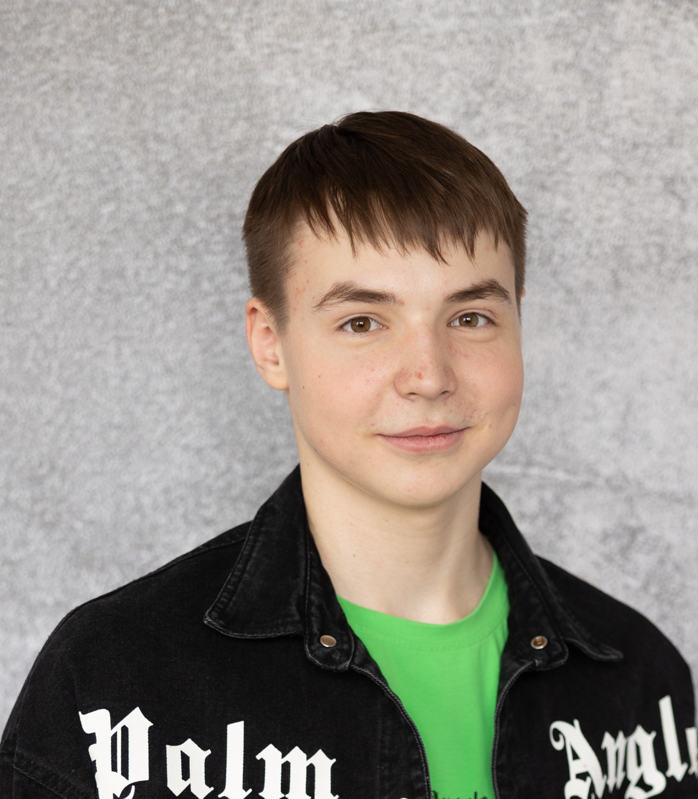 Teen boy with short brown hair, wearing a black jacket with white lettering over a bright green shirt, standing in front of a light grey textured background, smiling slightly at the camera. The image features AI Blemish Removal for a naturally clear and polished look.