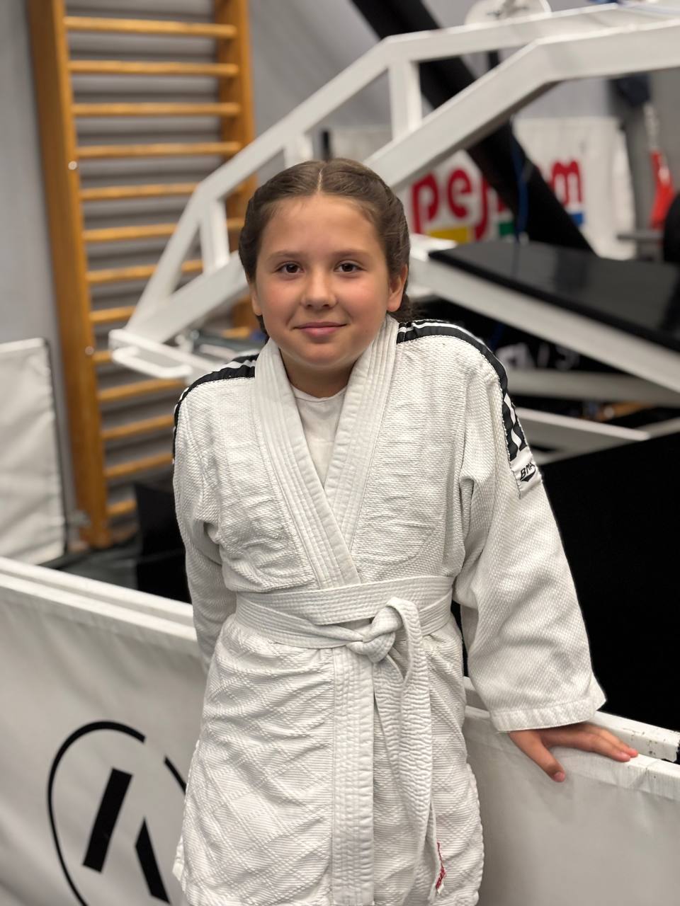 A young child wearing a white judo gi with a white belt stands indoors, smiling at the camera. Gym equipment and banners—one featuring “API Playground”—are visible in the blurred background.