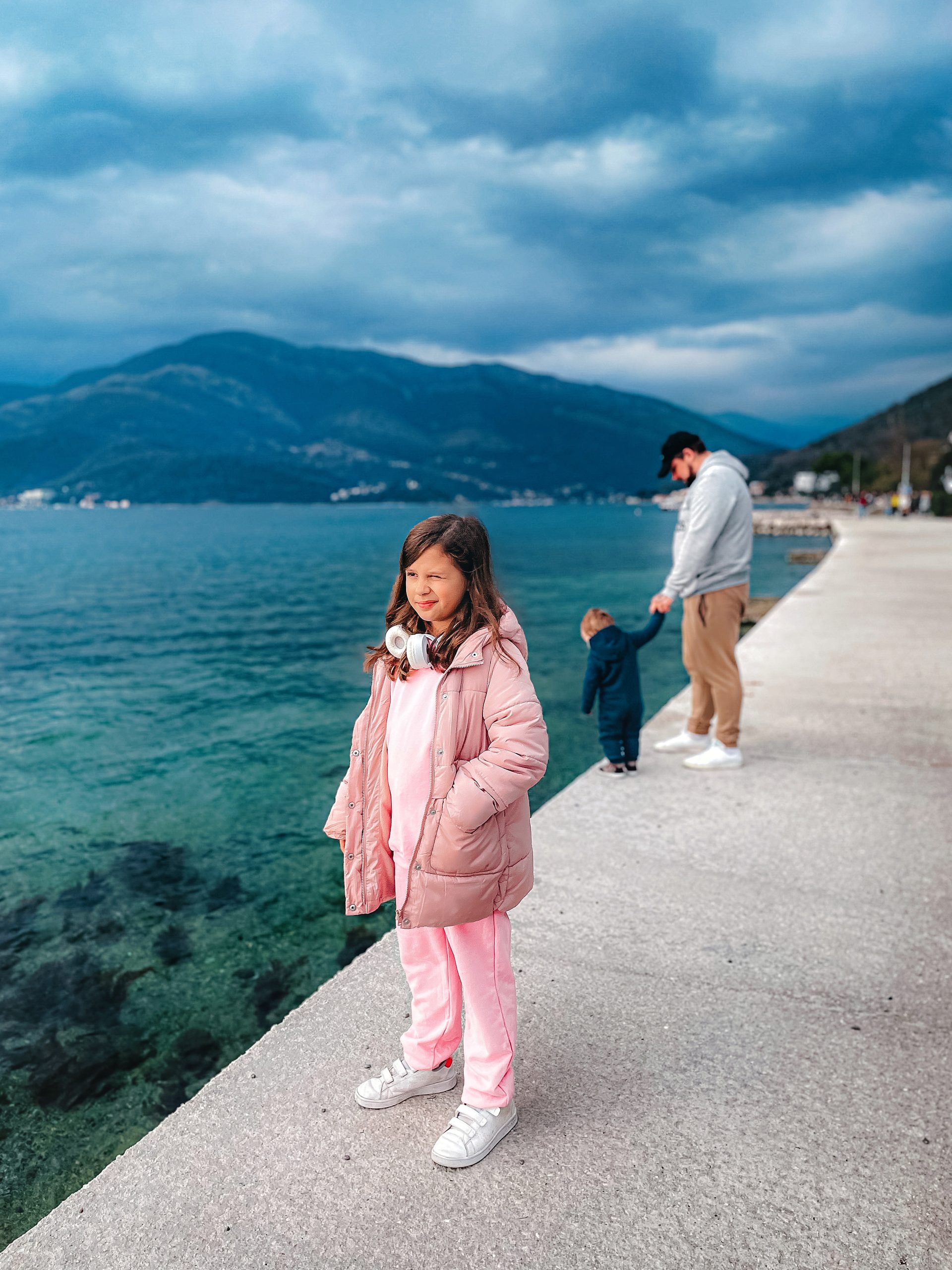 A young girl in a pink coat and headphones stands by a waterfront, much like an explorer in an API Playground, discovering new connections. In the background, a man and a child face the water, with mountains and cloudy skies visible across the bay.