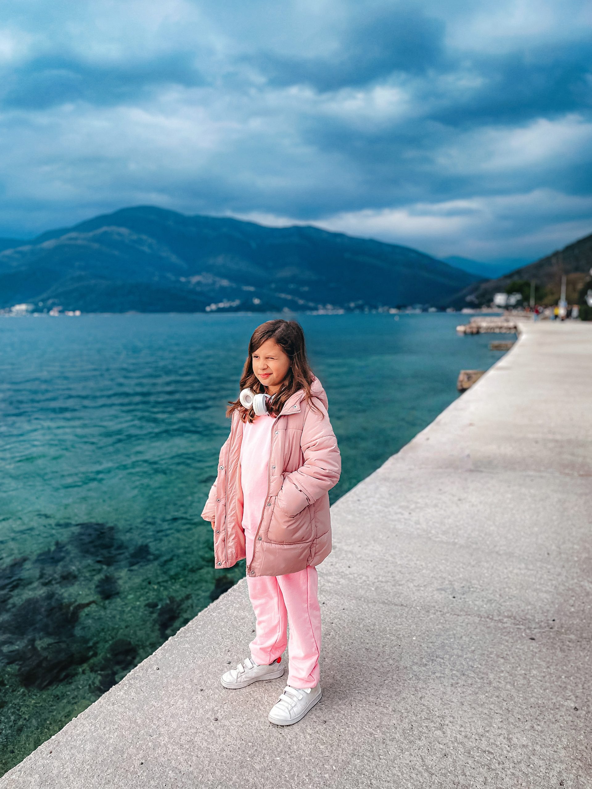 Young girl in a pink coat and trousers stands on a walkway by clear blue-green water, with mountains and cloudy skies in the background, evoking the sense of exploring an API Playground set within nature’s beauty.