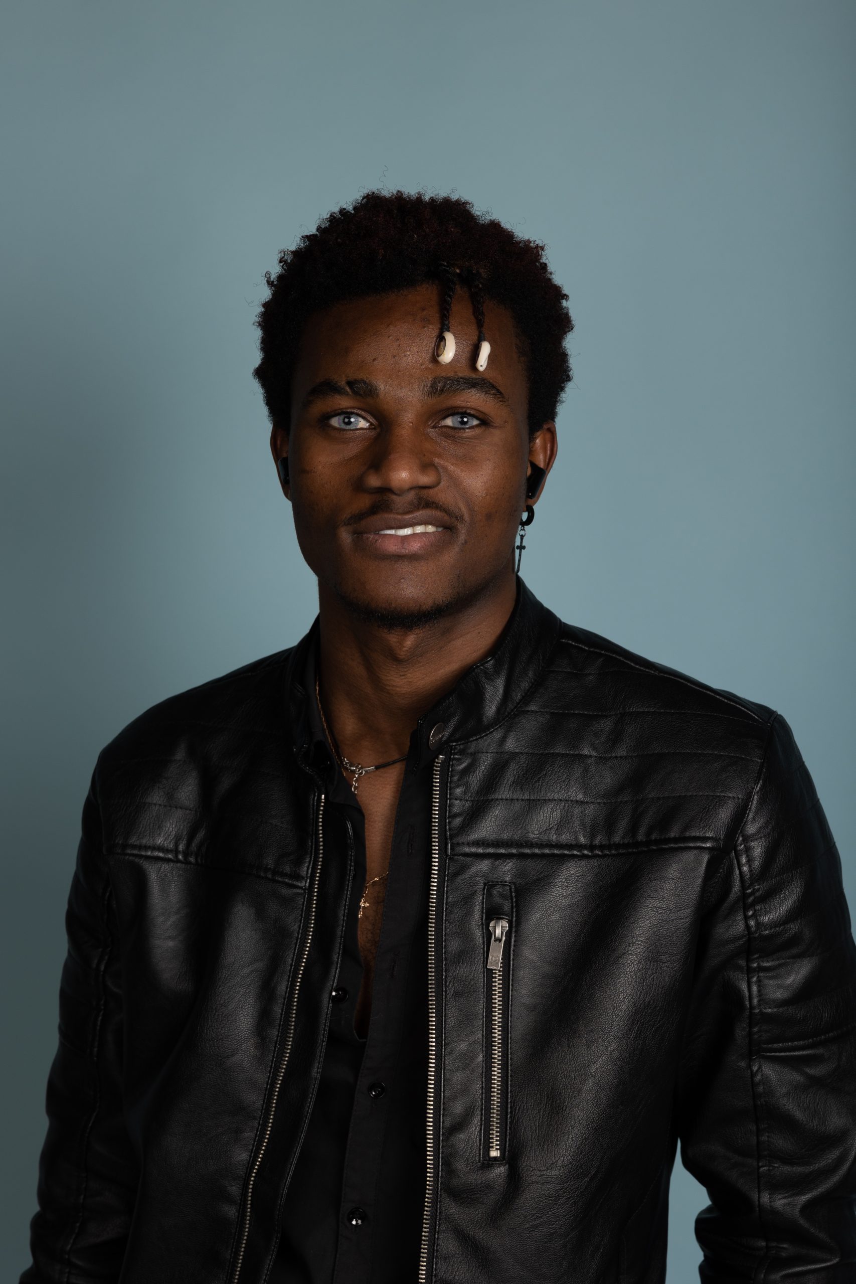 A man wearing a black leather jacket and black shirt stands in front of a plain blue background, smiling slightly. With short curly hair and two light beads plaited into the front, he exudes a confident presence that would fit right into the attractions industry.