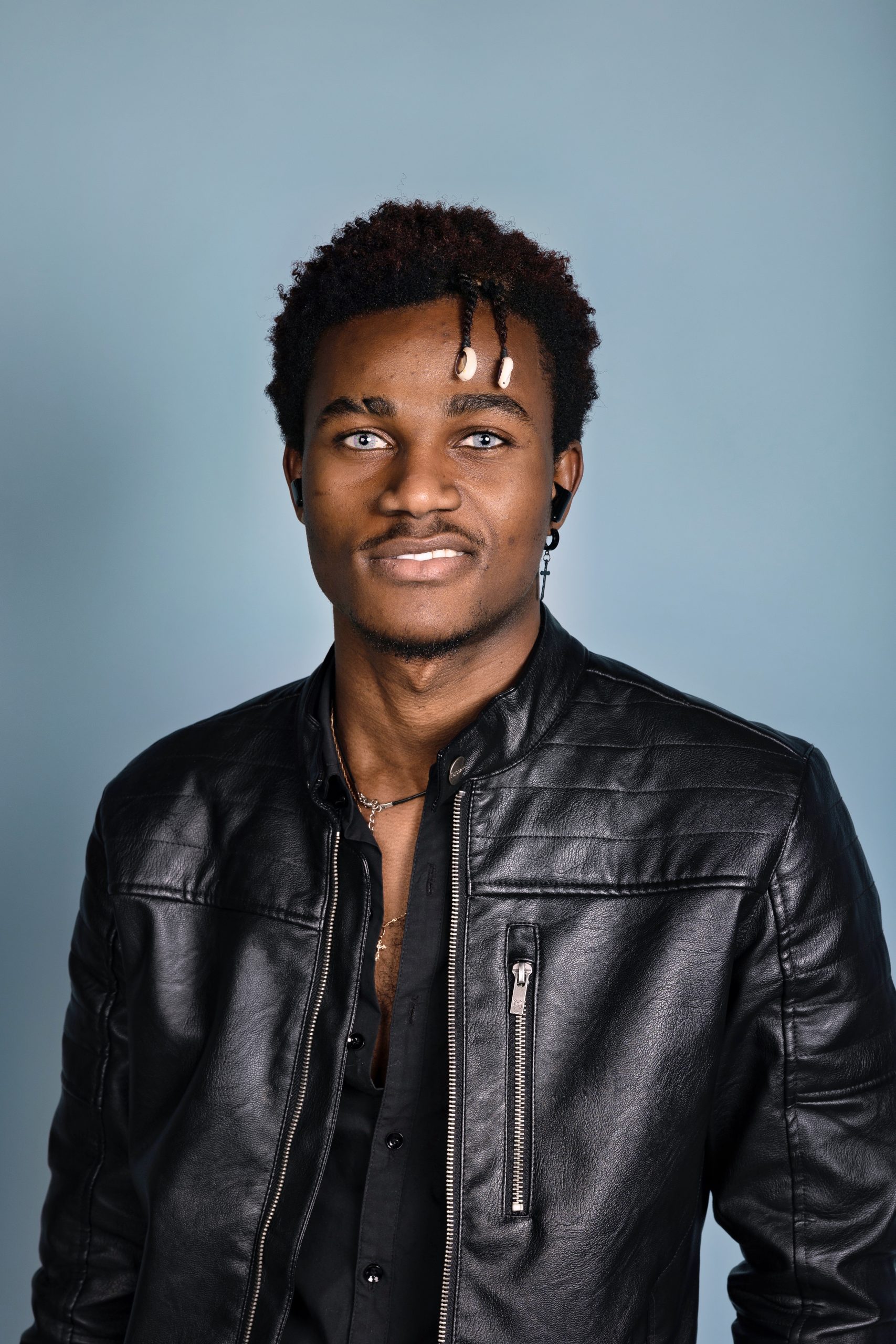 A young man with short curly hair, wearing a black leather jacket and a black shirt, stands in front of a light blue background, smiling slightly at the camera, embodying the confidence often seen among professionals in the attractions industry.