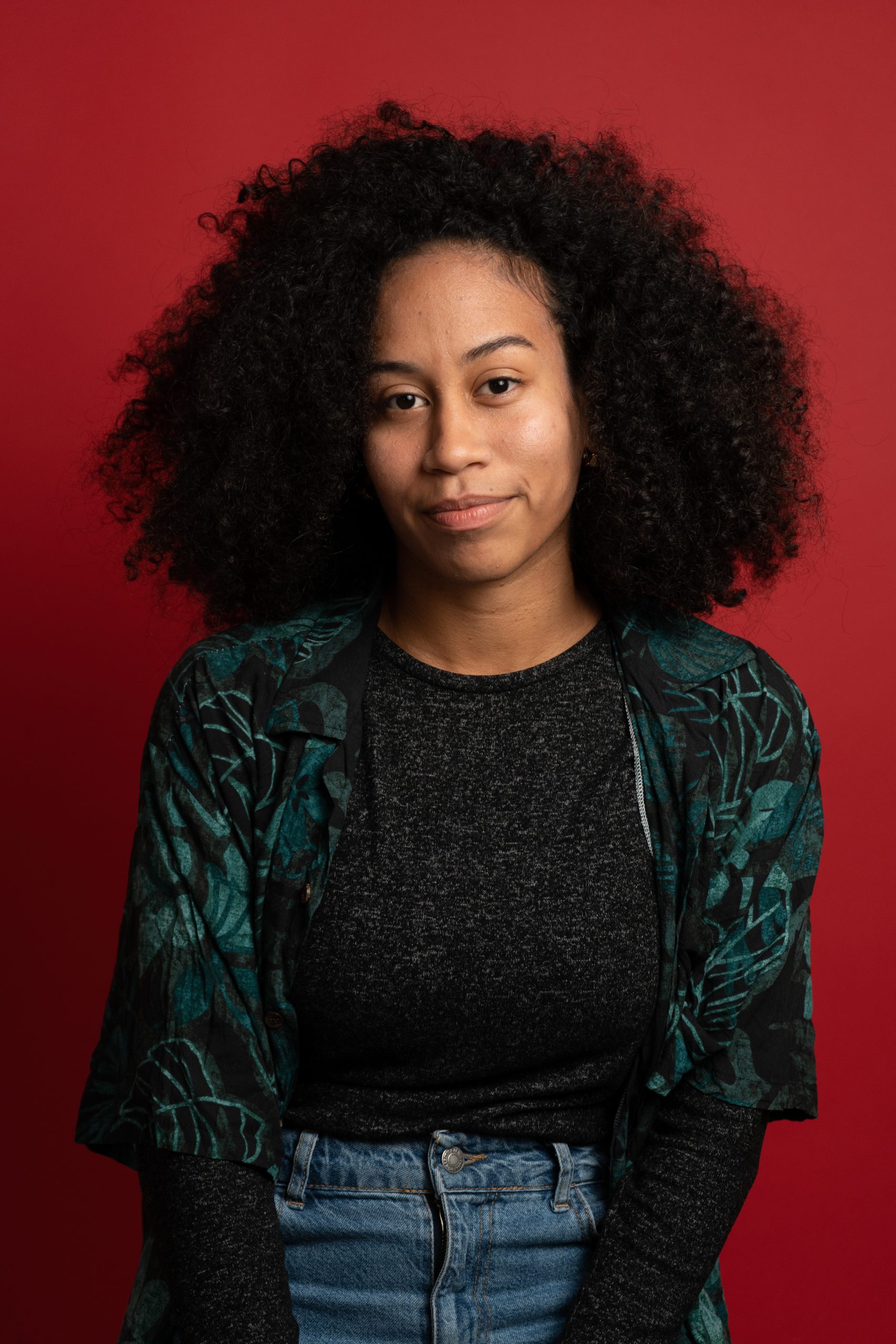 A woman with curly hair wearing a dark patterned shirt over a grey top and blue jeans sits in front of a solid red background, looking at the camera with a slight smile, as if she’s ready to explore the possibilities of an API Playground.