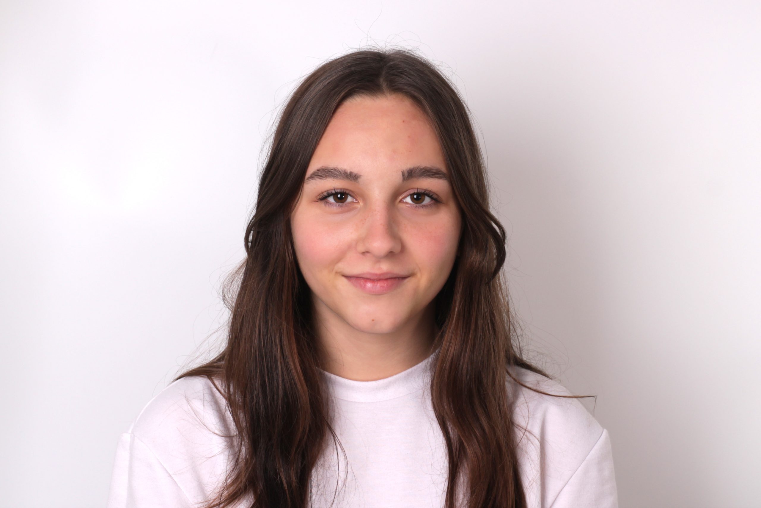 A young woman with long brown hair and a white shirt, her complexion enhanced by AI Blemish Removal, smiles softly in front of a plain white background.
